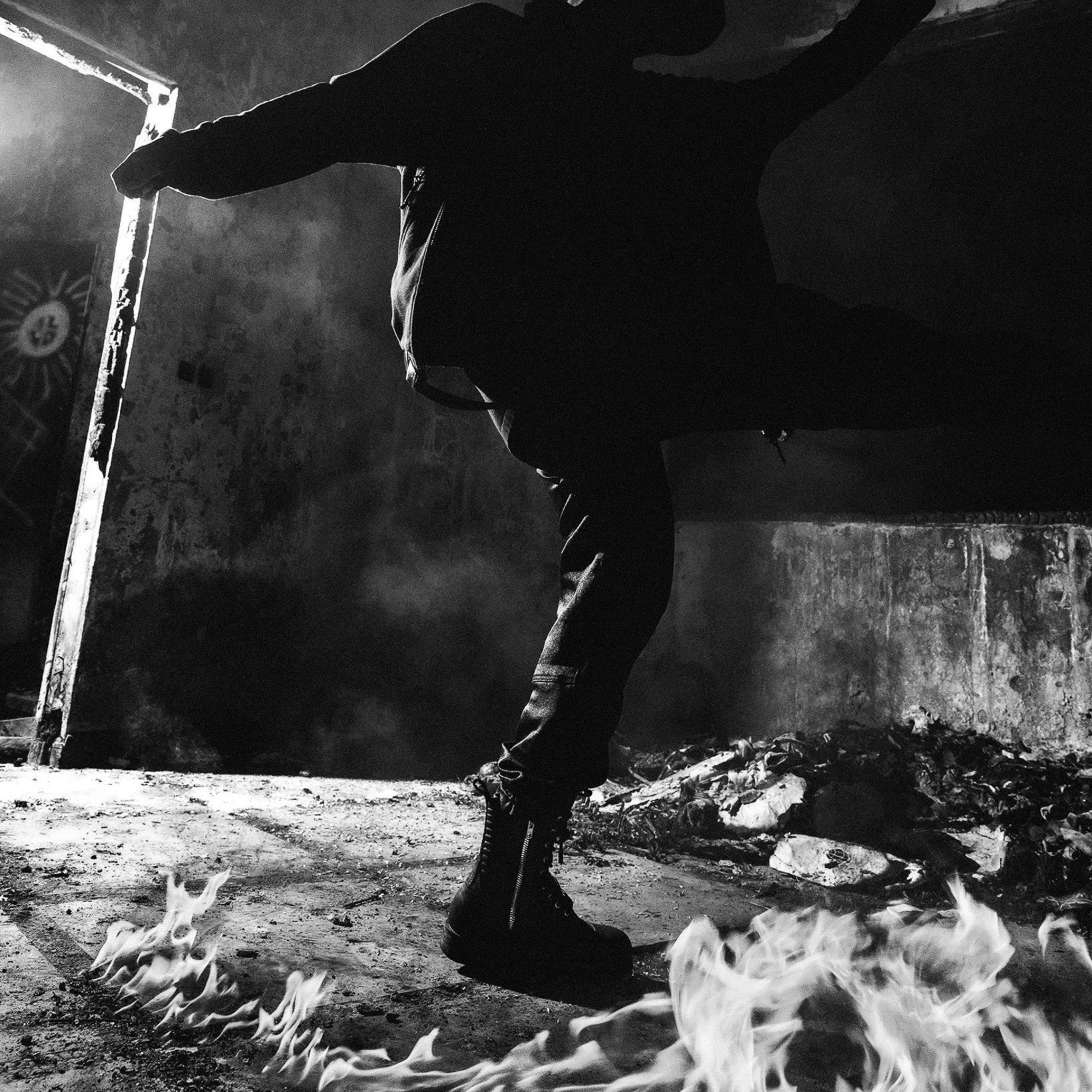 A dramatic black and white photograph of a person in a dark hoodie and boots, captured mid-motion with arms outstretched in a smoky, industrial interior with flames visible in the foreground.